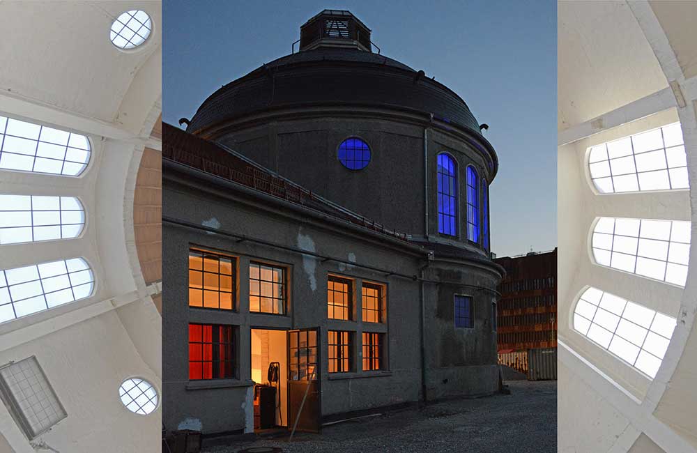 Gebäude mit kuppelartigem Dach, orange beleuchtete Fenster unten, blau beleuchtete Fenster oben bei Dämmerung.