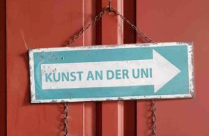 Schild mit Aufschrift Kunst an der Uni und Pfeil, hängend an einer roten Tür.