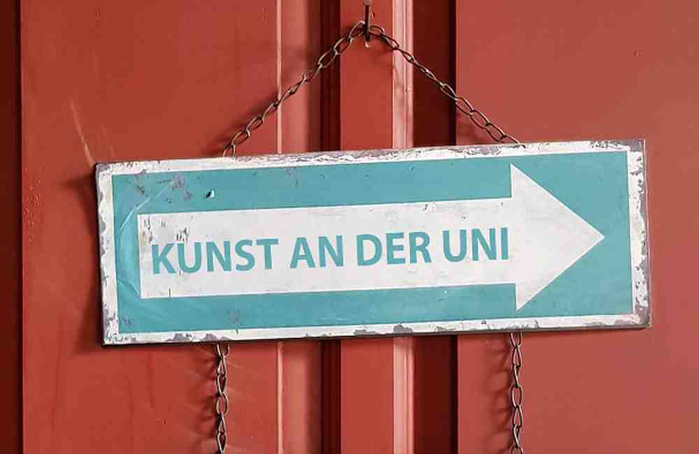 Schild mit Aufschrift Kunst an der Uni und Pfeil, hängend an einer roten Tür.
