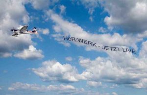Kleines Flugzeug zieht Werbebanner mit Schrift Hörwerk – Jetzt live vor wolkigem Himmel.