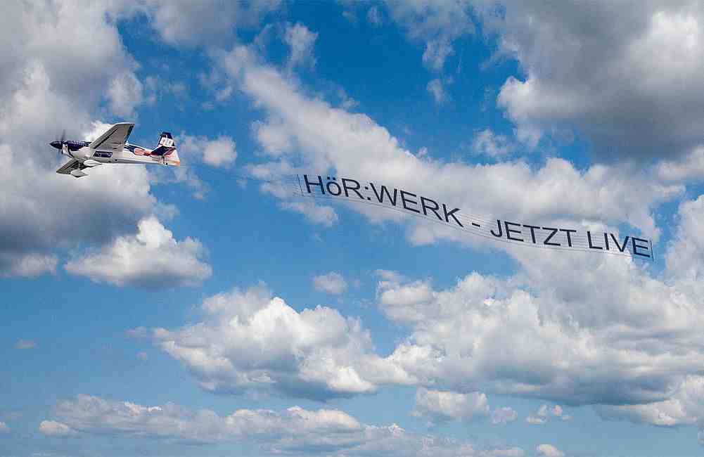 Kleines Flugzeug zieht Werbebanner mit Schrift Hörwerk – Jetzt live vor wolkigem Himmel.