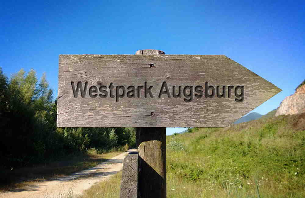 Holz-Wegweiser mit der Aufschrift Westpark Augsburg an einem ländlichen Pfad unter blauem Himmel.