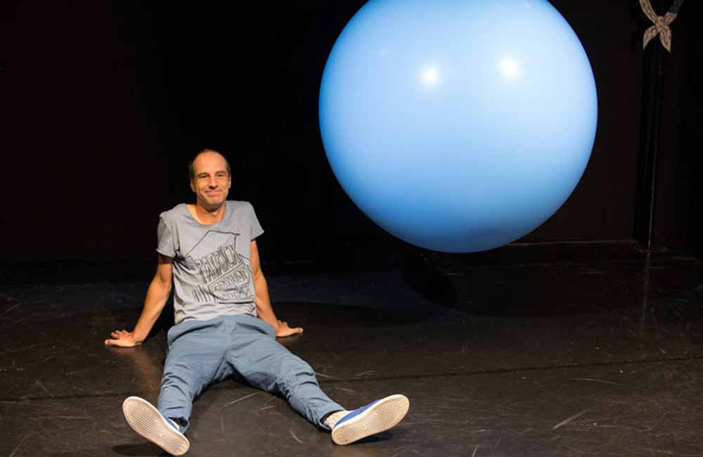 Mann sitzt auf dunklem Boden neben großem schwebendem blauen Ball.