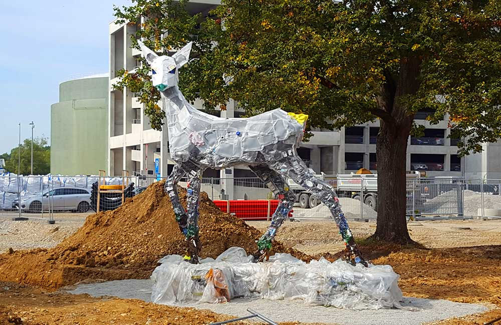 Skulptur eines Rehs aus recycelten Materialien auf einer Baustelle neben Baum und Erdwall.