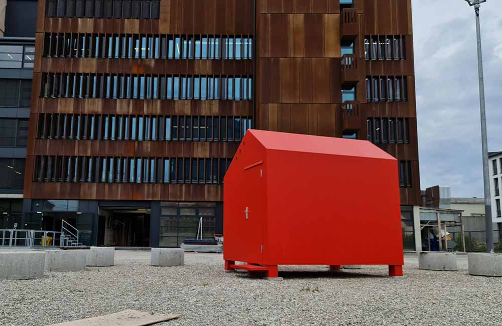 Kleines rotes Gebäude mit spitzem Dach auf kiesbedecktem Platz vor braunem Bürogebäude.