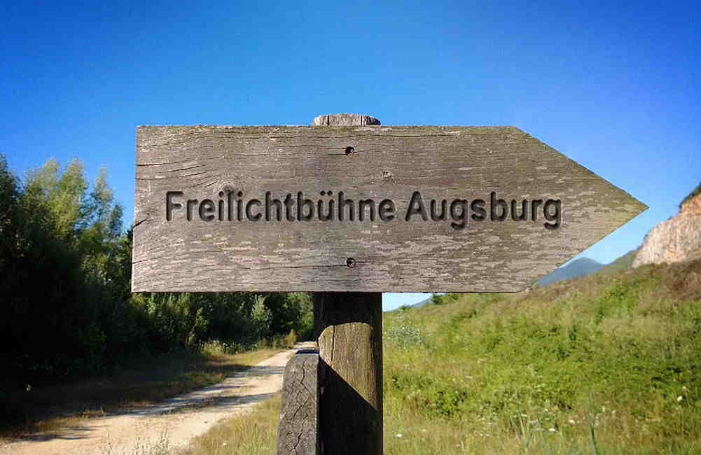 Holzwegweiser mit Aufschrift Freilichtbühne Augsburg zeigt auf Feldweg zwischen Bäumen und Gras.