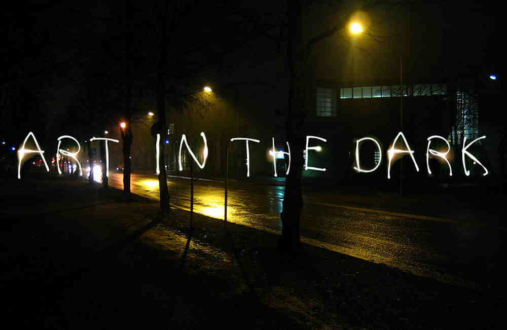Leuchtende Schrift ART IN THE DARK auf nasser Straße bei Nacht, beleuchtet von Laternen.