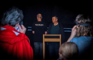 Personen in dunklem Raum, zwei Männer im Fokus, einer mit "ART IN THE DARK"-Shirt, weitere Personen im Vordergrund.