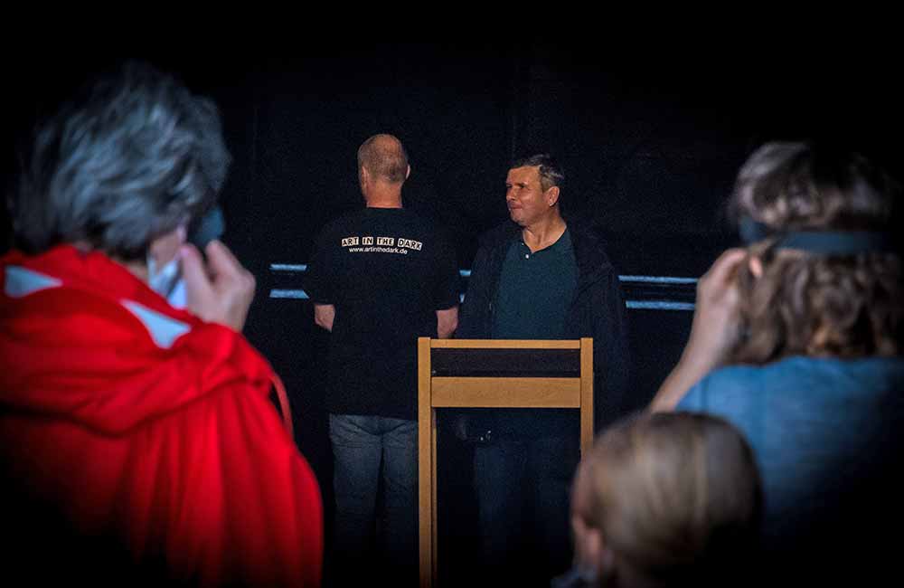 Personen in dunklem Raum, zwei Männer im Fokus, einer mit "ART IN THE DARK"-Shirt, weitere Personen im Vordergrund.
