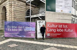 Zwei Bauzaunbanner mit Sprüchen zu Leben, Sport und Kultur vor historischem Gebäude, Mann geht vorbei.