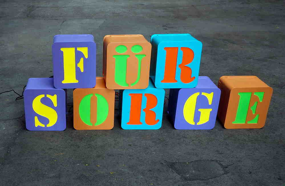 Mehrfarbige Buchstabenwürfel bilden das Wort FÜR SORGE auf grauem Boden.