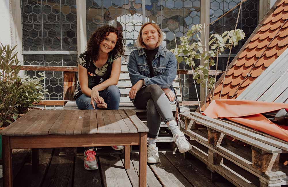 Zwei Frauen sitzen an Holztisch auf Terrasse mit Pflanzen und sechseckigen Fenstern im Hintergrund.