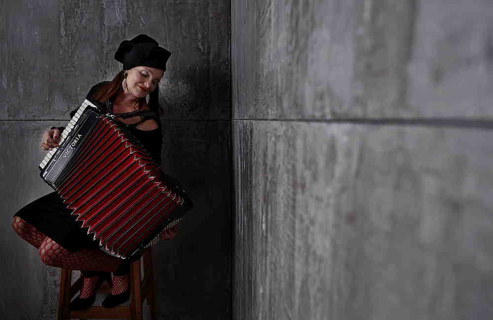 Frau in schwarzer Kleidung und Mütze sitzt auf Hocker und spielt rotes Akkordeon in grauem Raum.