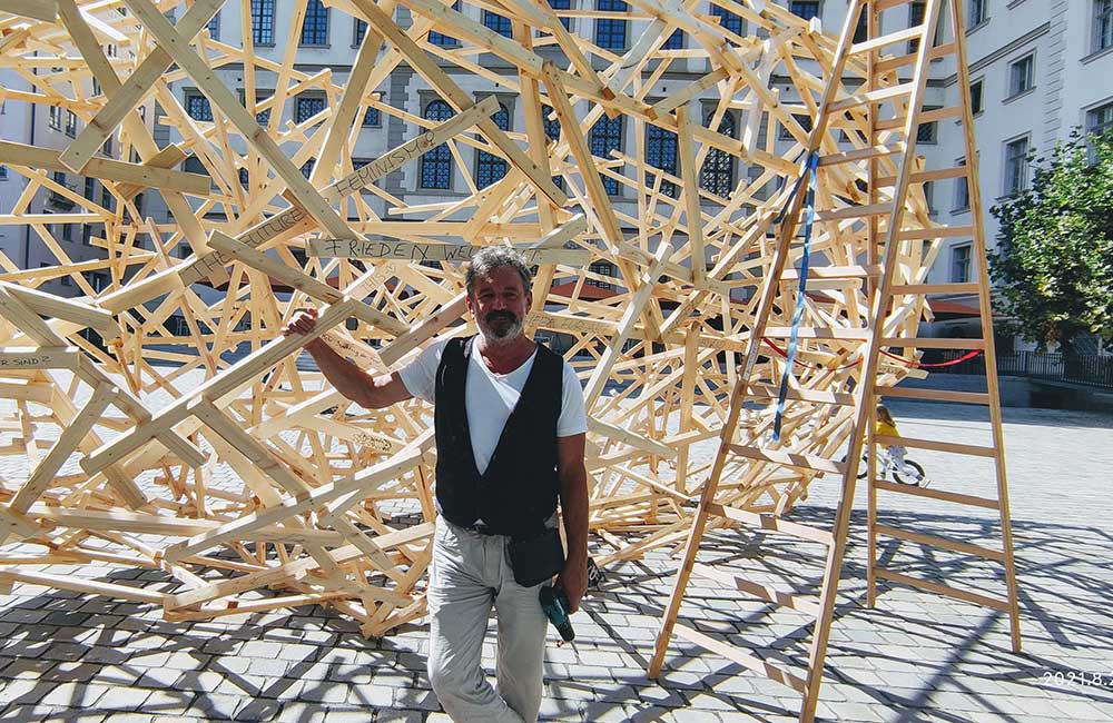 Mann steht vor komplexer Holzstruktur auf gepflastertem Platz vor historischem Gebäude.