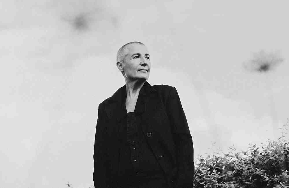 Ältere Person mit kurzgeschorenem Haar in dunklem Anzug vor hellem Himmel und Pflanzen.