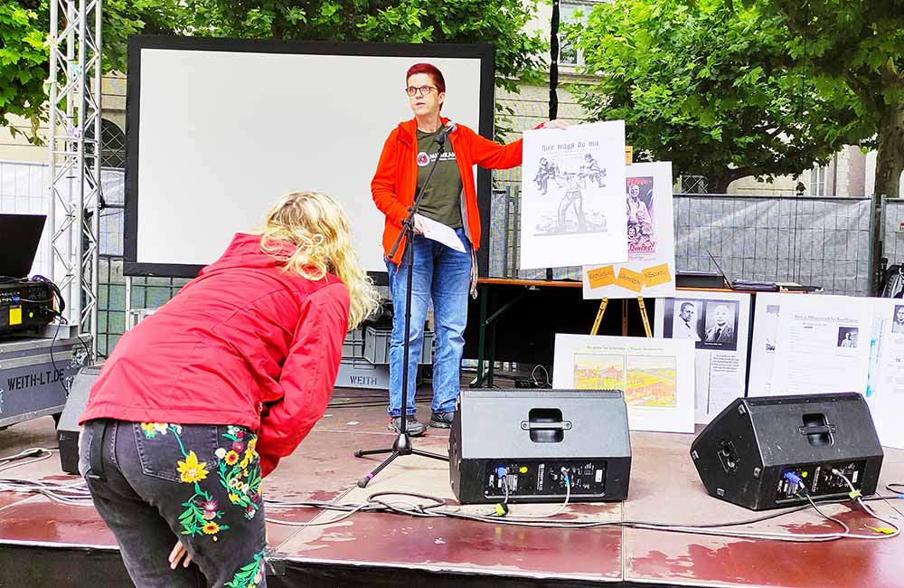 Person präsentiert auf Bühne ein Plakat vor Leinwand, weitere Plakate und Lautsprecher sichtbar.