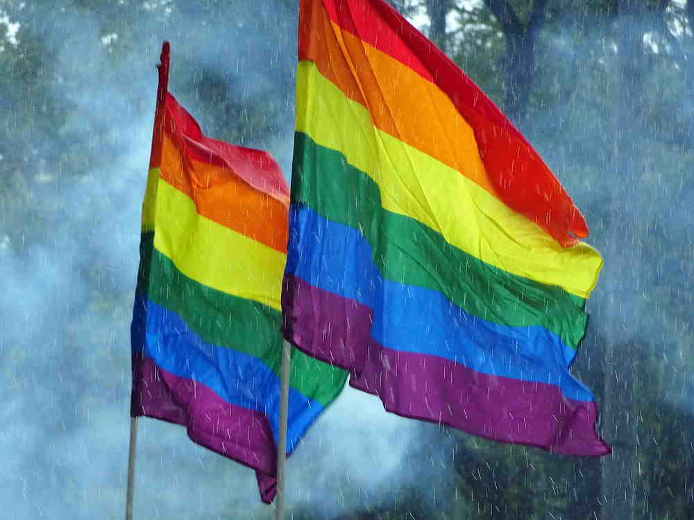Zwei wehende Regenbogenfahnen im Regen vor bewölktem Himmel.