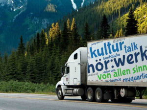 Weißer LKW mit beschriftetem Anhänger fährt an Nadelwald und Bergen vorbei.