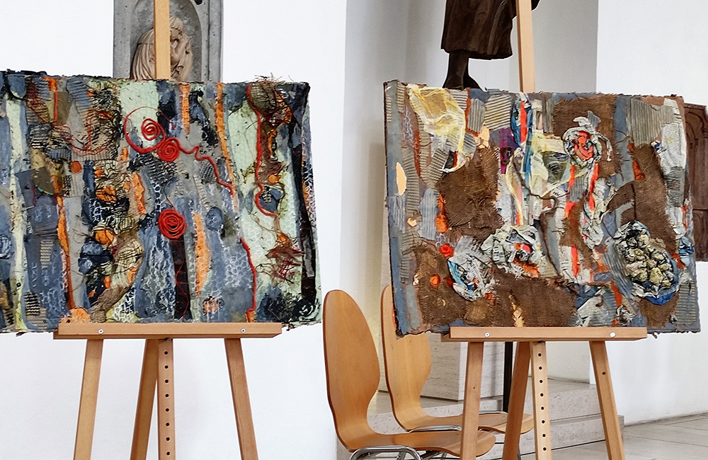 Abstrakte Gemälde mit verschiedenen Strukturen auf Staffeleien vor Holzkirchenstühlen und weißen Wänden.