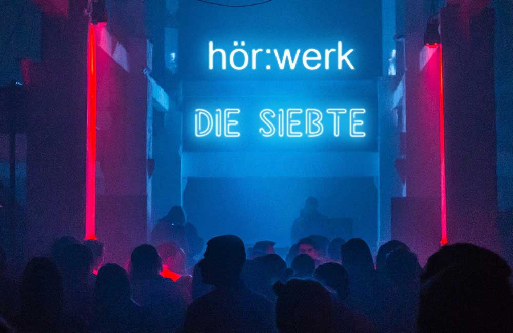 Menschenmenge bei Veranstaltung unter leuchtendem Schild hör werk DIE SIEBTE in blauem Licht