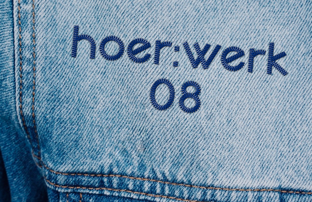 Blau gestickter Schriftzug hoer;werk 08 auf hellblauem Jeansstoff mit Naht.