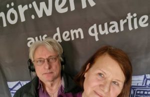 Zwei Personen vor einem Banner mit teilweisem Text in deutscher Sprache.