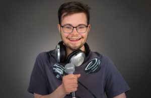 Junger Mann mit Brille, Kopfhörer um den Hals, hält Mikrofon vor grauem Hintergrund.