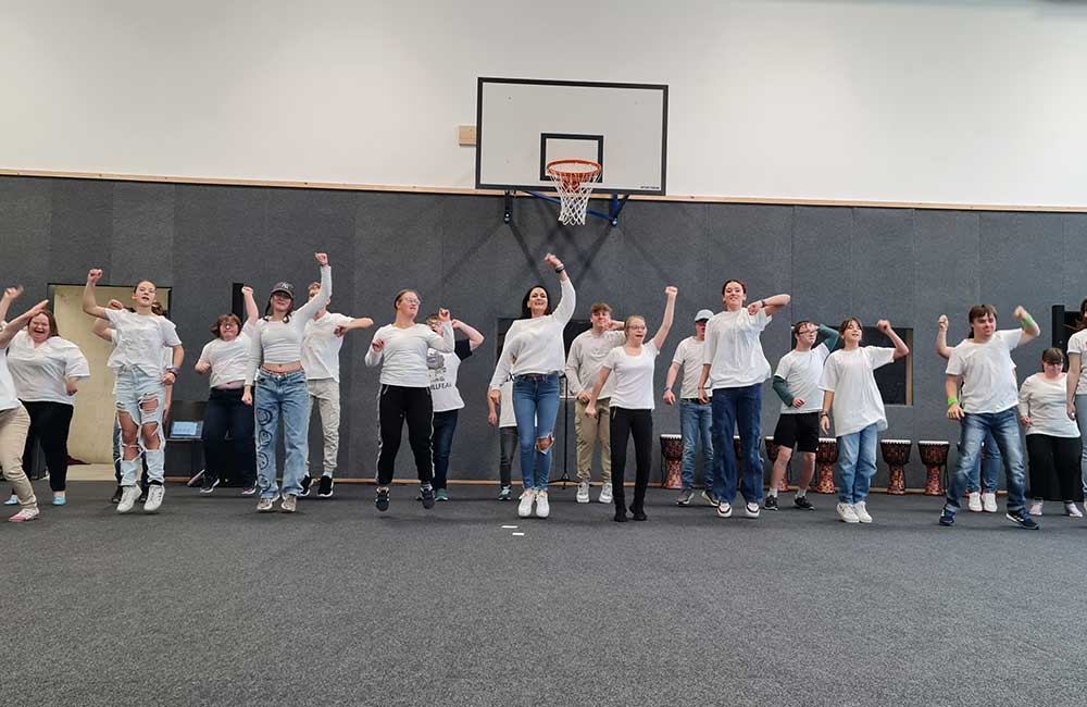 Gruppe von Menschen in weißen Oberteilen beim gemeinsamen Sport in einer Halle vor einem Basketballkorb.