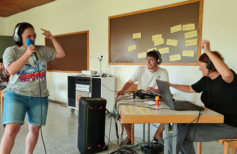 Drei Personen bei Musikaufnahme, eine singt mit Mikrofon, zwei bedienen Laptop und Mischpult.